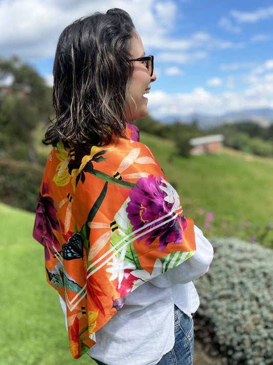 Modelo usando una pañoleta cuadrada grande amarrada como tipo chal, ilustrado fondo naranja vibrante con polinizadores colombianos, pañoletas cuadradas grandes para hacer top souvenir Colombia pañoleta turista, regalo original mujer pañoleta Colombia, pañoleta regalo navidad colombiana, pañoleta larga ilustrada Colombia. Ailé