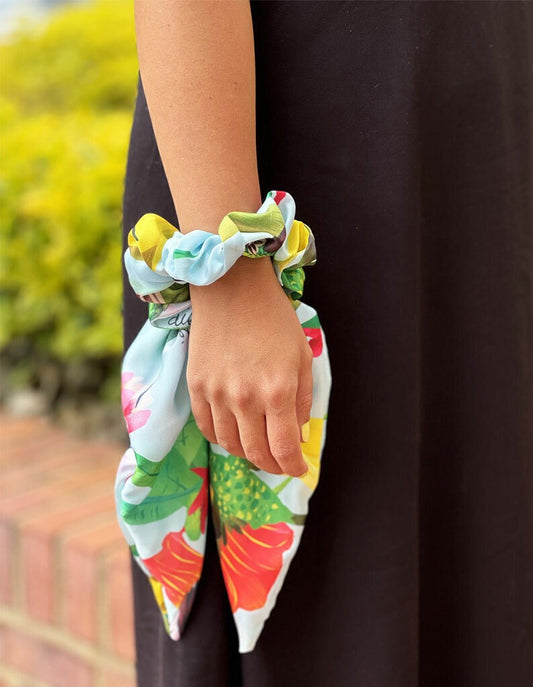 Scrunchie con lazo ilustrado ilustración "Colibrí Azul Colombia" — fondo celeste con colibríes en verde esmeralda, azul y amarillo, entre flores tropicales rojas y rosas. El colibrí es uno de los símbolos más icónicos de la biodiversidad colombiana regalo Día de la Madre bamba Colombia, scrunchie regalo Amor y Amistad Colombia, souvenir Colombia bamba turista, regalo original bamba Colombia, bamba regalo navidad colombiana, detalle empresarial bamba Colombia, regalo corporativo scrunchie colombiano. Ailé