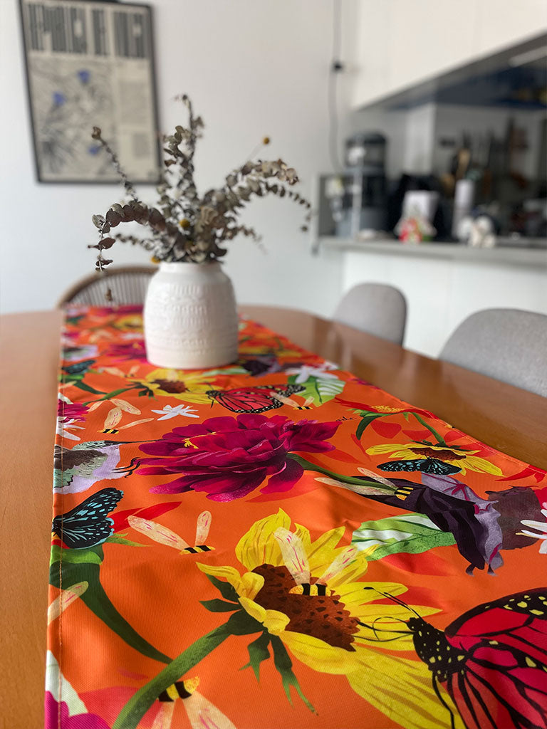 Camino de mesa ecológico ilustrado fondo naranja vibrante con polinizadores colombianos — mariposa monarca, colibrí, abejas, murciélago, flores grandes fucsia y girasoles amarillos, decoración colombiana para comedor, Colombian design table runner gift, recycled fabric table runner Colombia, hecho en Colombia, camino de mesa con diseño de fauna colombiana. Ailé