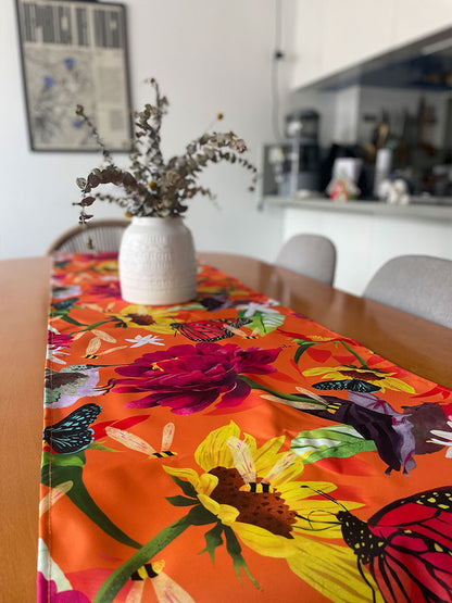 Camino de mesa ecológico ilustrado fondo naranja vibrante con polinizadores colombianos — mariposa monarca, colibrí, abejas, murciélago, flores grandes fucsia y girasoles amarillos, decoración colombiana para comedor, Colombian design table runner gift, recycled fabric table runner Colombia, hecho en Colombia, camino de mesa con diseño de fauna colombiana. Ailé