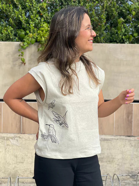 Mujer usando camiseta Ailé con ilustración de colibríes, moda inspirada en aves de Colombia, camiseta ilustración línea negra, camiseta boceto colibrí, camiseta beige estampada mujer, camiseta dibujo minimalista aves, camiseta arte lineal Colombia, Colombian hummingbird t-shirt, hummingbird print shirt handmade, Colombia wildlife clothing gift, birdwatcher gift Colombia shirt, illustrated hummingbird apparel. Ailé