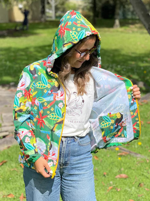 Mujer con chaqueta rompevientos ilustrada fondo verde oscuro con tucanes colombianos entre flores tropicales rojas, chaqueta impermeable diseño colombiano, cortavientos con estampado de fauna colombiana, colombian eco jacket illustrated, colombian wildlife waterproof jacket, colombian fauna windbreaker gift, Colombian toucan gift. Ailé