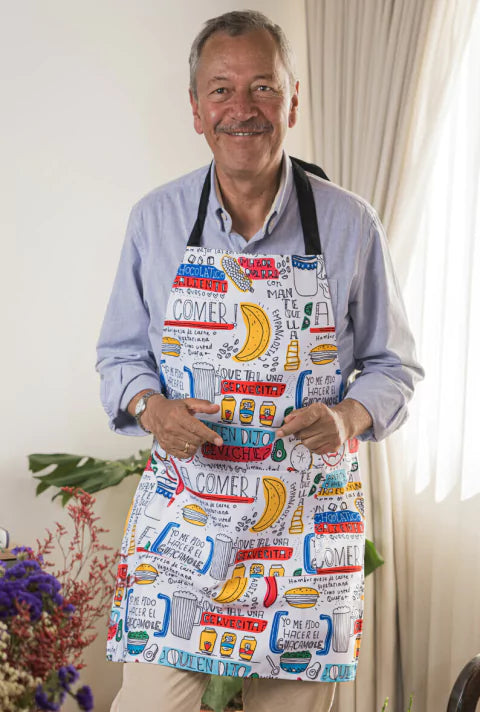 Delantal con diseño de dichos y frases colombianas de comida — textos ilustrados: "¡A comer!", "¿Quién dijo ceviche?", "¿Qué tal una cervecita?", "Yo me pido hacer el guacamole", "Chocolatico caliente con queso" — acompañados de ilustraciones de empanadas, cervezas, aguacates, dichos colombianos, mandil, frases típicas de Colombia,regalo de cocina colombiano, delantal ilustrado, colombianos en el exterior y turistas. Ailé